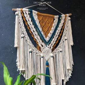 May include: A large macrame wall hanging with a geometric design in white, mustard yellow, and teal blue. The hanging has a fringe border and is made with cotton cord.