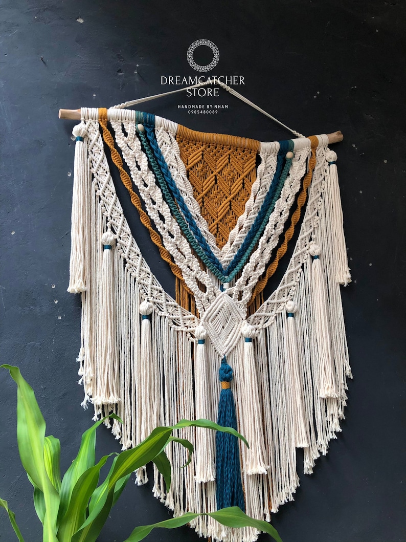 May include: A large macrame wall hanging with a geometric design in white, mustard yellow, and teal blue. The hanging has a fringe border and is made with cotton cord.
