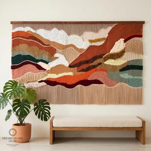 May include: A large, textured wall hanging with a landscape design in shades of brown, orange, green, and cream. The artwork is mounted on a wooden frame and features a variety of textures, including woven and knotted elements. The bottom has long fringe.