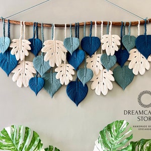May include: A wall hanging made of macrame with a variety of blue and white leaves. The leaves are made of cotton cord and are hanging from a wooden branch. The wall hanging is labelled "Dreamcatcher Store" and "Handmade by Nham 0905480089".