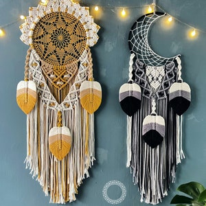 May include: Two handmade dreamcatchers hanging on a blue wall. The dreamcatcher on the left is a circular design with a yellow, white, and brown color scheme. The dreamcatcher on the right is a crescent moon shape with a black, white, and gray color scheme. Both dreamcatchers have feathers hanging from the bottom.