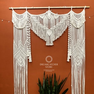 May include: A white macrame wall hanging with a geometric design. The hanging is made of cotton cord and has a fringe at the bottom. The hanging is attached to a wooden dowel rod. The text "Dreamcatcher Store" is visible in the image. Handmade by Nham.