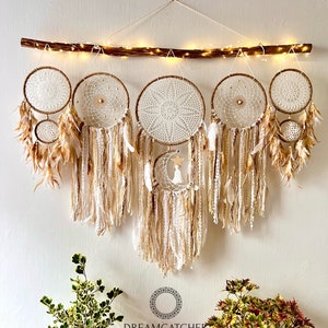 May include: A set of six beige and white dreamcatchers with feathers hanging from a wooden branch. The dreamcatchers are arranged in a row and have different designs. The dreamcatchers are decorated with beads and other embellishments. The text "Dreamcatcher Store" and "Handmade by NHAM 0905480089" is at the bottom of the image.