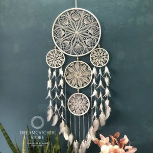Op de afbeelding: Witte gehaakte droomvanger met vier hoepels en witte veren die eronder hangen. De droomvanger hangt aan een teal muur. De tekst "Dreamcatcher Store Handmade by Nhan 0905480089" staat onderaan de afbeelding.