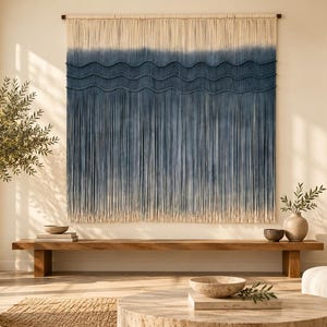 May include: A large rectangular macrame wall hanging featuring a cream-colored top section that transitions into a deep blue ombre gradient. The design includes three horizontal wavy cord patterns layered over long, vertical fringe.