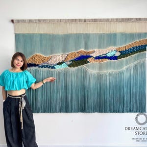 May include: Large, textured wall hanging with a landscape design. The artwork features a gradient of blue and beige tones, with a central mountain-like form in shades of brown, green, and blue. The bottom has long, vertical fringe. The top has a wooden rod.
