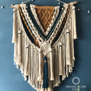 Puede incluir: Un gran tapiz de macramé tejido de forma intrincada con un patrón geométrico en tonos beige, amarillo mostaza y azul verdoso. El tapiz presenta una variedad de nudos y borlas, y está terminado con una varilla de madera en la parte superior.