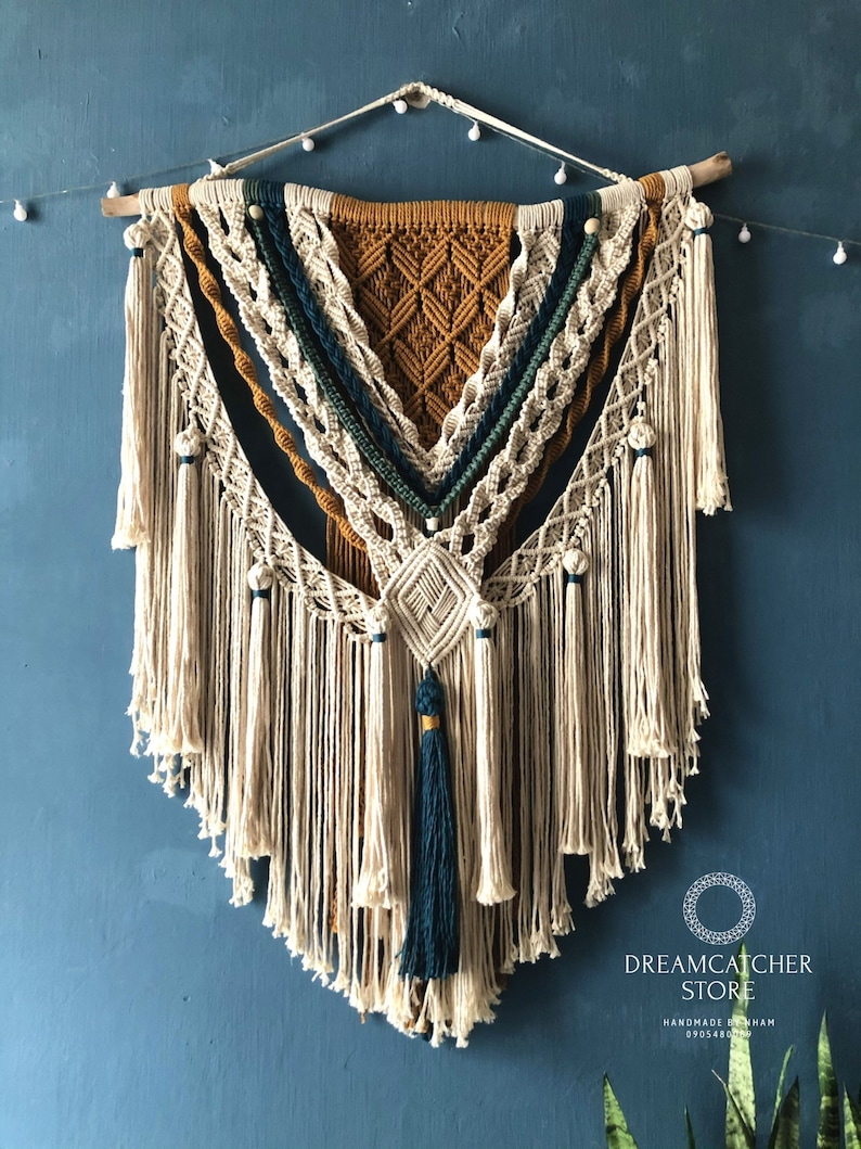 May include: A large, intricately woven macrame wall hanging with a geometric pattern in shades of beige, mustard yellow, and teal. The hanging features a variety of knots and tassels, and is finished with a wooden dowel at the top.
