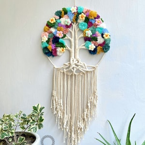Pode incluir: Uma tapeçaria de macramé de parede colorida com um design de árvore da vida com pompons em tons de rosa, azul, verde, amarelo e roxo. A tapeçaria é feita com cordão de algodão natural e tem uma franja na parte inferior. O texto "Dreamcatcher Store" é visível na parte inferior da imagem.