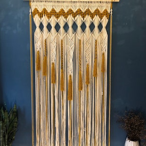 May include: A macrame wall hanging with a white and gold color scheme. The hanging features a geometric pattern with tassels at the bottom.