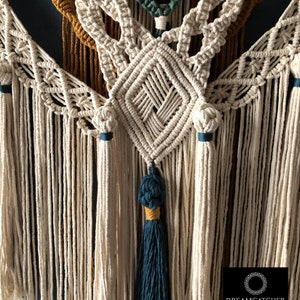 May include: A close-up of a macrame wall hanging with a diamond pattern in the center. The hanging is made with natural colored cord and has teal and mustard yellow accents. The hanging has a teal tassel at the bottom.