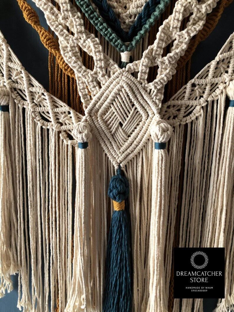 May include: A close-up of a macrame wall hanging with a diamond pattern in the center. The hanging is made with natural colored cord and has teal and mustard yellow accents. The hanging has a teal tassel at the bottom.