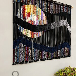 May include: A colorful macrame wall hanging with a black and blue wave design. The hanging is made with a variety of colorful cords and is attached to a wooden dowel.