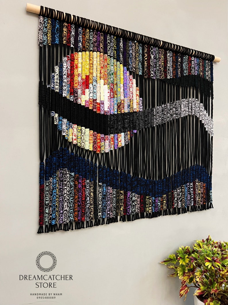 May include: A colorful macrame wall hanging with a black and blue wave design. The hanging is made with a variety of colorful cords and is attached to a wooden dowel.