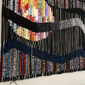 May include: A colorful macrame wall hanging with a black and white wave design. The hanging is made with a variety of colored cords and is attached to a wooden dowel.