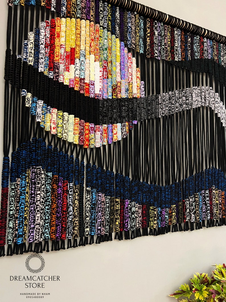 May include: A colorful macrame wall hanging with a black and white wave design. The hanging is made with a variety of colored cords and is attached to a wooden dowel.