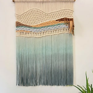 May include: A macrame wall hanging with a gradient of blue and green colors. The hanging features a woven design with a brown and orange stripe.