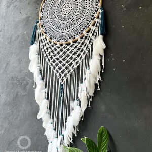 May include: White and teal dreamcatcher with white feathers and beads hanging from a brown hoop. The dreamcatcher is hanging on a black wall with a green plant in the bottom right corner. The text "Dreamcatcher Store" and "Handmade by Nham 0905480089" is at the bottom left corner of the image.
