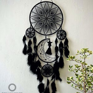May include: Black dreamcatcher with a crescent moon and star design, feathers, and beads. The dreamcatcher is hanging on a white wall with a small green plant in the background. The text "DREAMCATCHER STORE" and "HANDMADE BY NHAM" is at the bottom of the image.