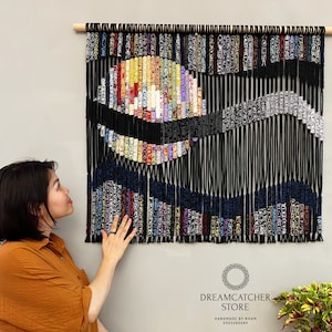 May include: A colorful abstract wall hanging made of yarn and string. The hanging features a large circle in the center with a wave-like pattern below it. The hanging is black with colorful accents. The text "Dreamcatcher Store" is visible at the bottom of the image.