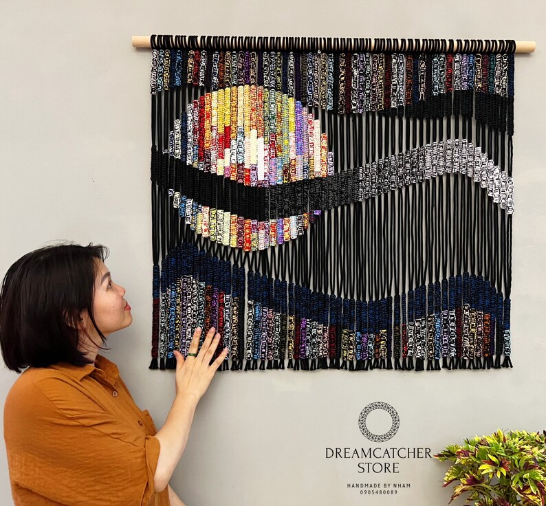 May include: A colorful abstract wall hanging made of yarn and string. The hanging features a large circle in the center with a wave-like pattern below it. The hanging is black with colorful accents. The text "Dreamcatcher Store" is visible at the bottom of the image.
