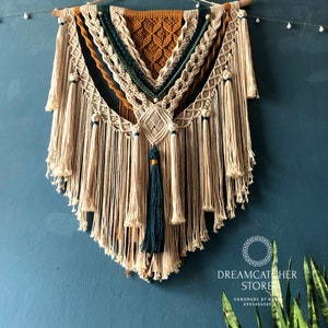 May include: A large macrame wall hanging with a geometric design in shades of beige, brown, and teal. The hanging features a variety of knots and tassels, and is finished with a wooden dowel at the top. The hanging is displayed against a teal wall, with a plant in the bottom right corner. The text "Dreamcatcher Store" is visible in the bottom right corner of the image.
