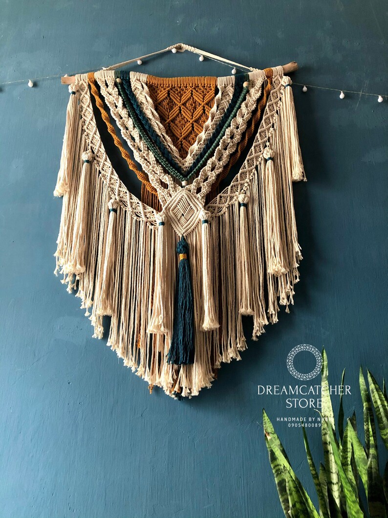 May include: A large macrame wall hanging with a geometric design in shades of beige, brown, and teal. The hanging features a variety of knots and tassels, and is finished with a wooden dowel at the top. The hanging is displayed against a teal wall, with a plant in the bottom right corner. The text "Dreamcatcher Store" is visible in the bottom right corner of the image.