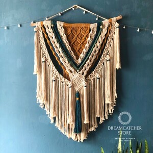 May include: A large macrame wall hanging with a geometric pattern in shades of beige, mustard yellow, and teal. The hanging features a variety of knots and tassels, and is adorned with wooden beads. The hanging is labeled "Dreamcatcher Store Handmade by Nham 0948087".