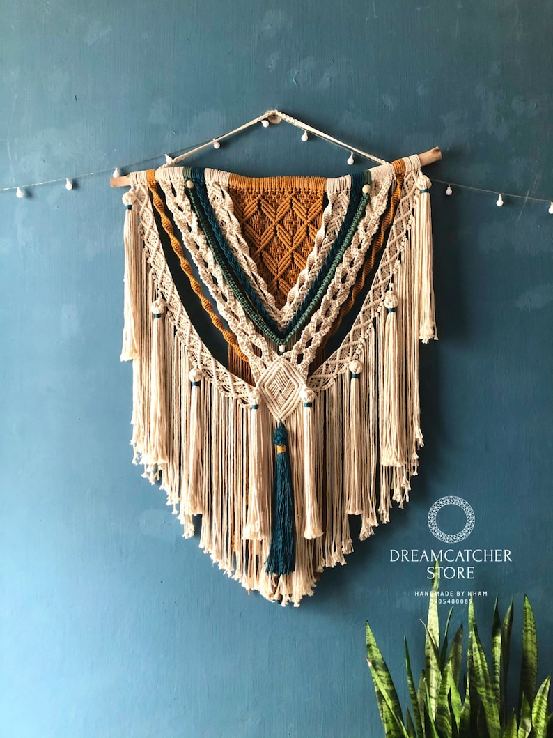 May include: A large macrame wall hanging with a geometric pattern in shades of beige, mustard yellow, and teal. The hanging features a variety of knots and tassels, and is adorned with wooden beads. The hanging is labeled "Dreamcatcher Store Handmade by Nham 0948087".