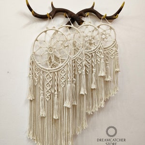 May include: Three white macrame dreamcatchers hanging from a brown antler wall decor. The dreamcatchers have a circular design with a web-like pattern and long, flowing fringe. The text "Dreamcatcher Store Handmade by Nham 0905480889" is at the bottom of the image.