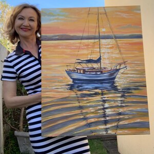 May include: A woman holds a painting of a sailboat on a calm body of water at sunset. The sky is a vibrant orange and yellow, with a hint of pink. The water is a deep blue, reflecting the colors of the sky. The sailboat is white with a blue sail, and it is anchored in the middle of the painting.