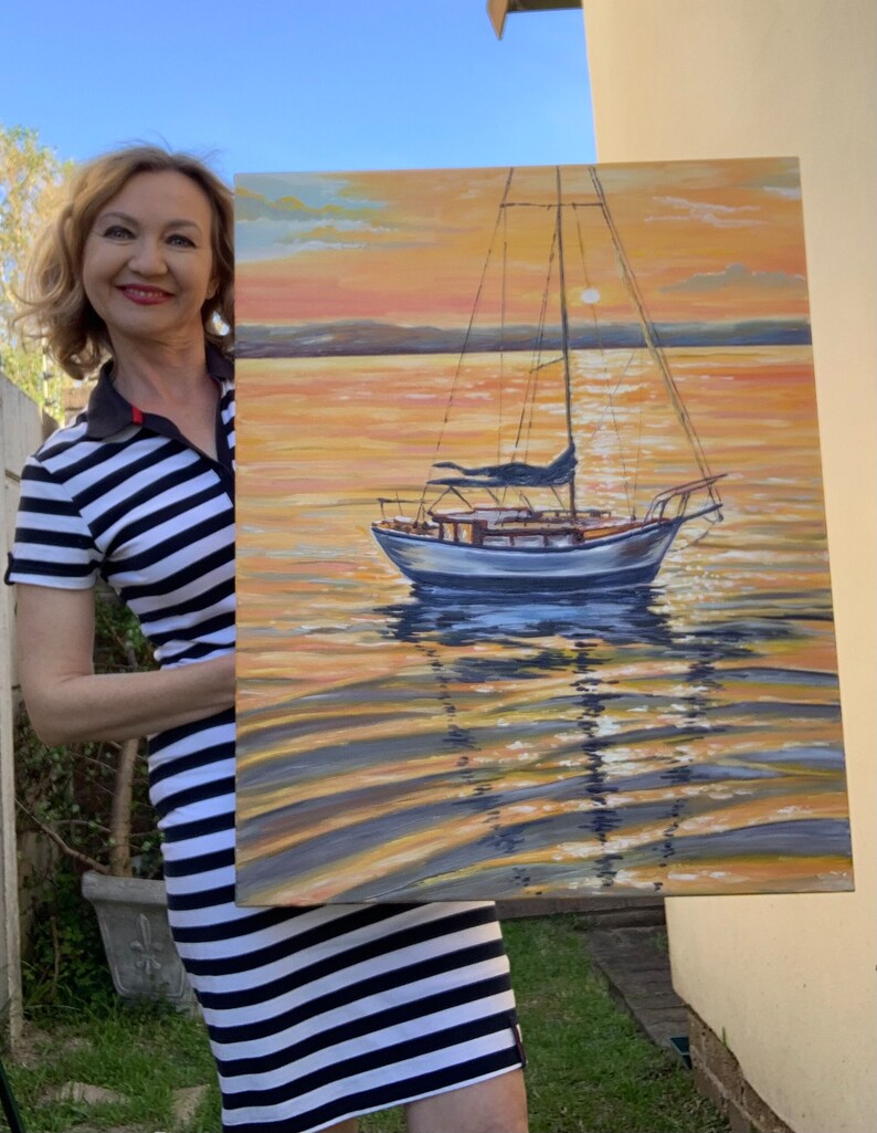 May include: A woman holds a painting of a sailboat on a calm body of water at sunset. The sky is a vibrant orange and yellow, with a hint of pink. The water is a deep blue, reflecting the colors of the sky. The sailboat is white with a blue sail, and it is anchored in the middle of the painting.
