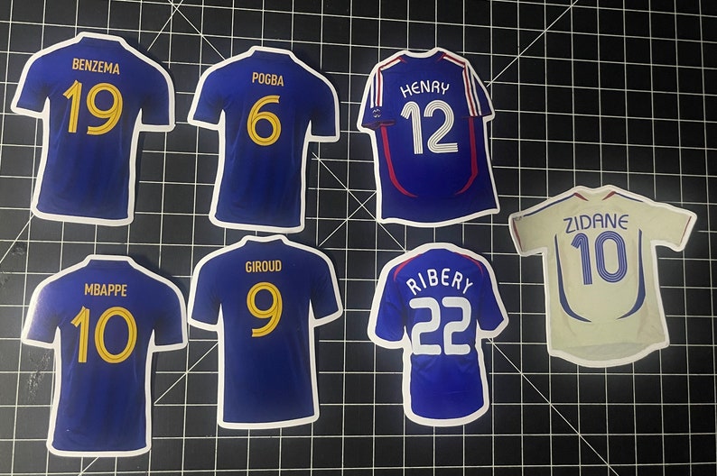 May include: A set of 8 blue and white soccer jersey stickers with the names and numbers of famous French soccer players. The names include Benzema, Pogba, Henry, Zidane, Mbappe, Giroud, Ribery.