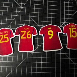May include: Four red soccer jerseys with yellow numbers on the back. The jerseys have the names "Ansu Fati 25", "Pedri 26", "Gavi 9", and "Ramos 15" printed on the back.