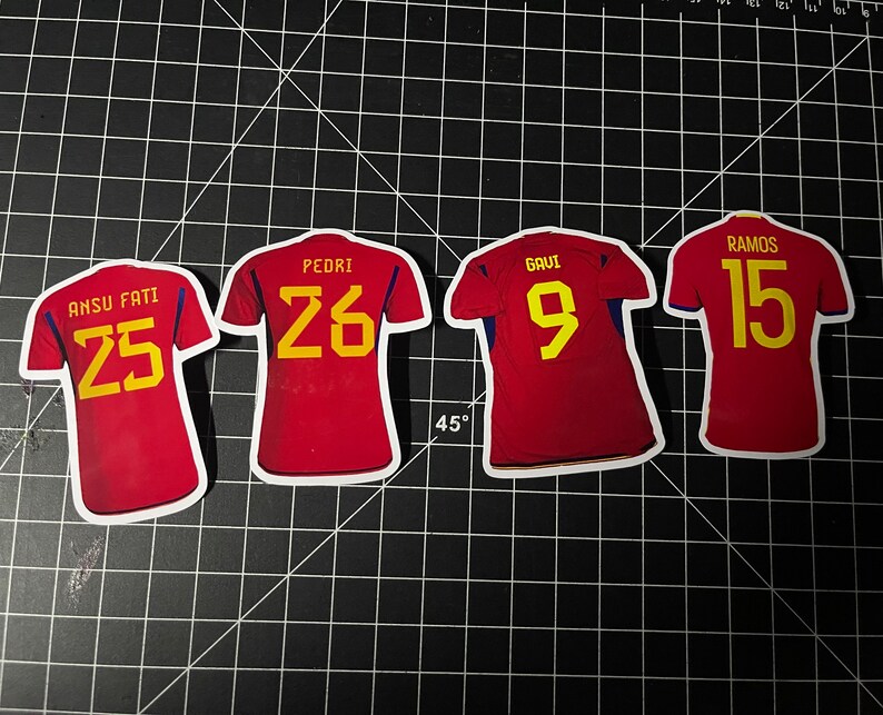 May include: Four red soccer jerseys with yellow numbers on the back. The jerseys have the names "Ansu Fati 25", "Pedri 26", "Gavi 9", and "Ramos 15" printed on the back.