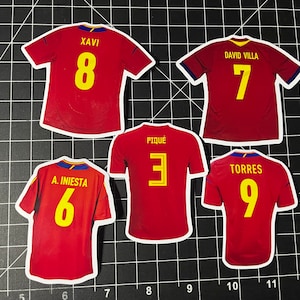 May include: Five red soccer jerseys with yellow numbers. The jerseys have the names "Xavi", "David Villa", "A. Iniesta", "Piqu&eacute;", and "Torres" on the back.