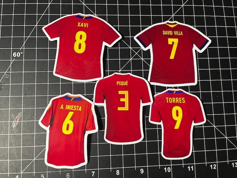 May include: Five red soccer jerseys with yellow numbers. The jerseys have the names "Xavi", "David Villa", "A. Iniesta", "Piqu&eacute;", and "Torres" on the back.