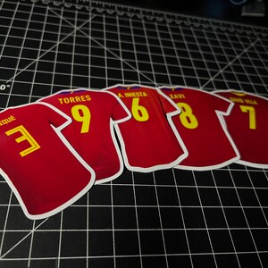 May include: Five red soccer jerseys with yellow numbers and white lettering. The jerseys have the names "Pique", "Torres", "A. Iniesta", "Xavi", and "David Villa" on the back.