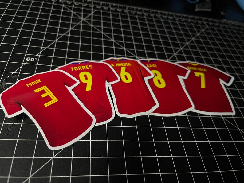 May include: Five red soccer jerseys with yellow numbers and white lettering. The jerseys have the names "Pique", "Torres", "A. Iniesta", "Xavi", and "David Villa" on the back.