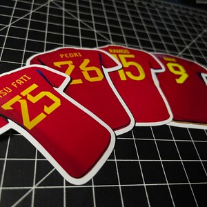 May include: Four red soccer jerseys with yellow numbers and player names. The jerseys have the numbers 25, 26, 15, and 9.