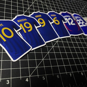 May include: A set of blue soccer jerseys with gold numbers. The jerseys have the names of famous soccer players on the back, including Mbappe, Benzema, Giroud, Pogba, Henry, and Zidane.
