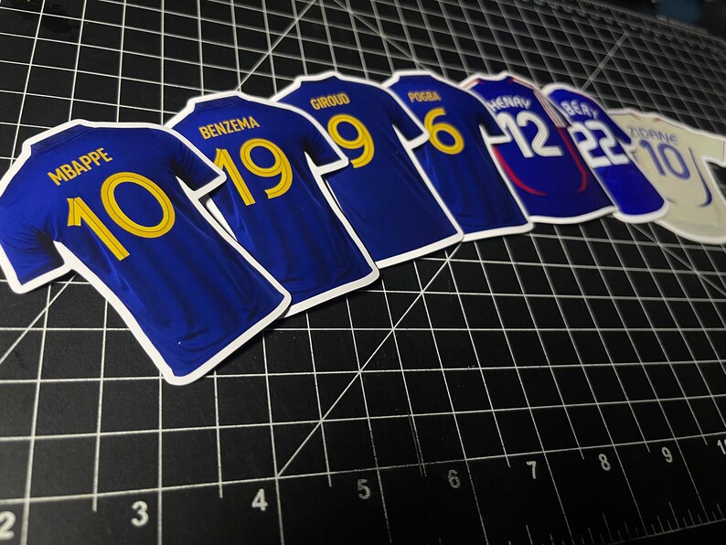 May include: A set of blue soccer jerseys with gold numbers. The jerseys have the names of famous soccer players on the back, including Mbappe, Benzema, Giroud, Pogba, Henry, and Zidane.