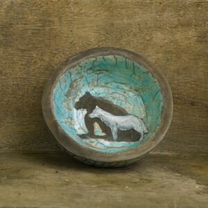 Könnte beinhalten: Eine runde Keramikschale mit einem dunkelbraunen Rand und einem türkisfarbenen Inneren mit rissiger Glasur. In der Mitte der Schale befindet sich eine gemalte Illustration eines dunkelbraunen Bären, der hinter einem kleineren, weißen Tier steht.