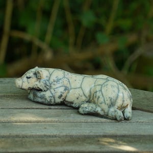 Könnte beinhalten: Eine kleine, cremefarbene Keramikfigur eines ruhenden Bären. Die Oberfläche weist ein ausgeprägtes, dunkelgraues Craquelé-Glasurmuster auf, das die Form des Bären nachzeichnet und den Kopf, die gefalteten Gliedmaßen und den Rücken hervorhebt.