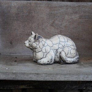 Puede incluir: Una figura de gato de cerámica con un patrón agrietado, similar a un mosaico, en blanco y negro. El gato está en posición de descanso, con la cola enrollada. La figura está colocada sobre una superficie de madera, con una figura de dragón tallada visible.