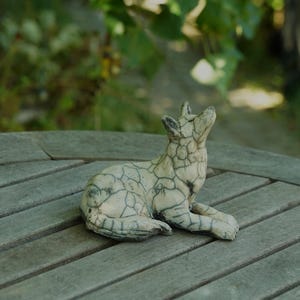Może przedstawiać: Ceramiczna figurka wilka w pozycji siedzącej, wyjącego. Rzeźba ma złamany biały kolor podstawowy z wyraźnym, ciemnoszarym wzorem spękań szkliwa pokrywającym całą powierzchnię, co podkreśla kontury ciała zwierzęcia i jego anatomiczne detale.