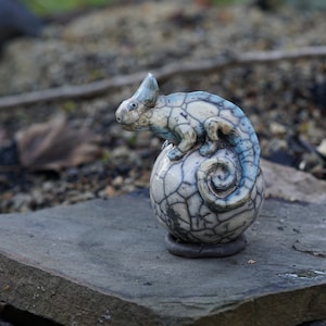Könnte beinhalten: Eine Keramik-Chamäleon-Skulptur mit blauem und weißem Glasur, die auf einer runden, rissigen Keramik-Kugel sitzt.