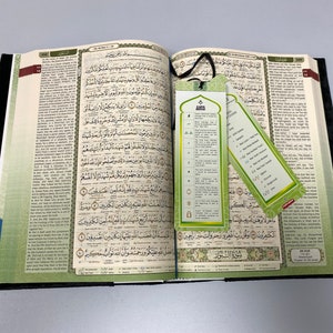 May include: An open Quran with two green bookmarks. The bookmarks feature Arabic text and symbols. The Quran is bound in black leather with gold accents.