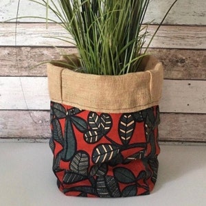 Peut inclure: Un pot de fleurs en tissu rouge et vert avec un dessus en toile de jute. Le pot est rempli de plantes vertes ressemblant à de l'herbe.