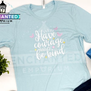 May include: A light blue t-shirt with the text "Have courage and be kind" in white with pink butterflies and stars. The shirt is laid flat on a white surface with a pair of colorful sneakers, blue jeans, and a blue glitter baseball cap.